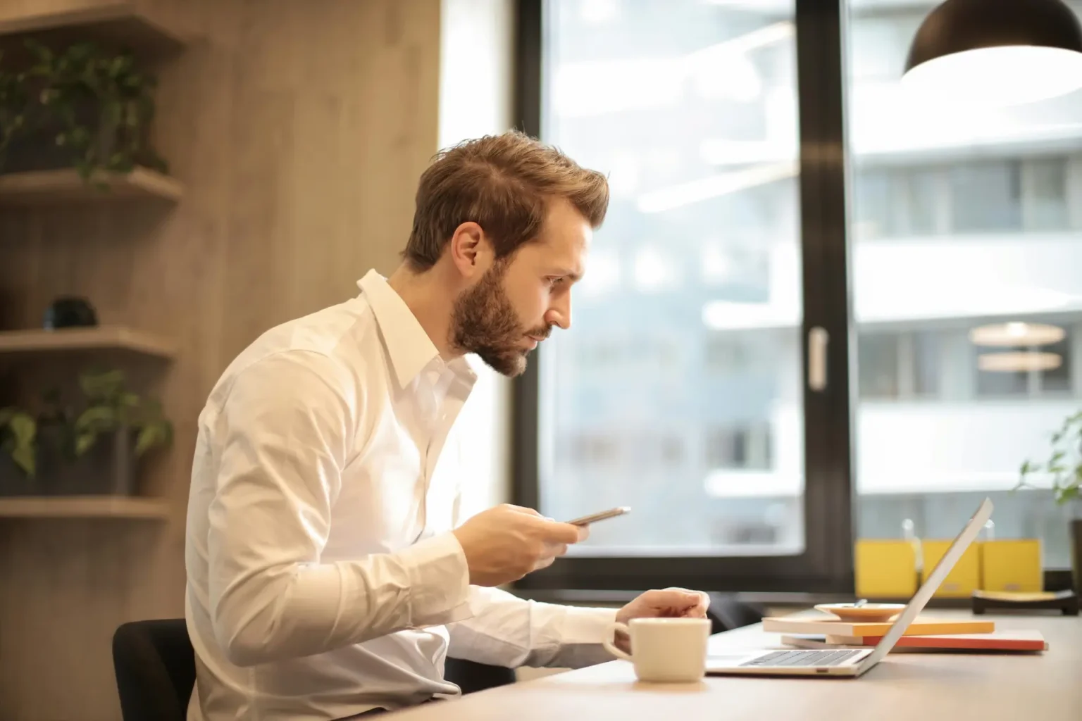Male at a desk looking at his laptop while also scrolling on a mobile phone